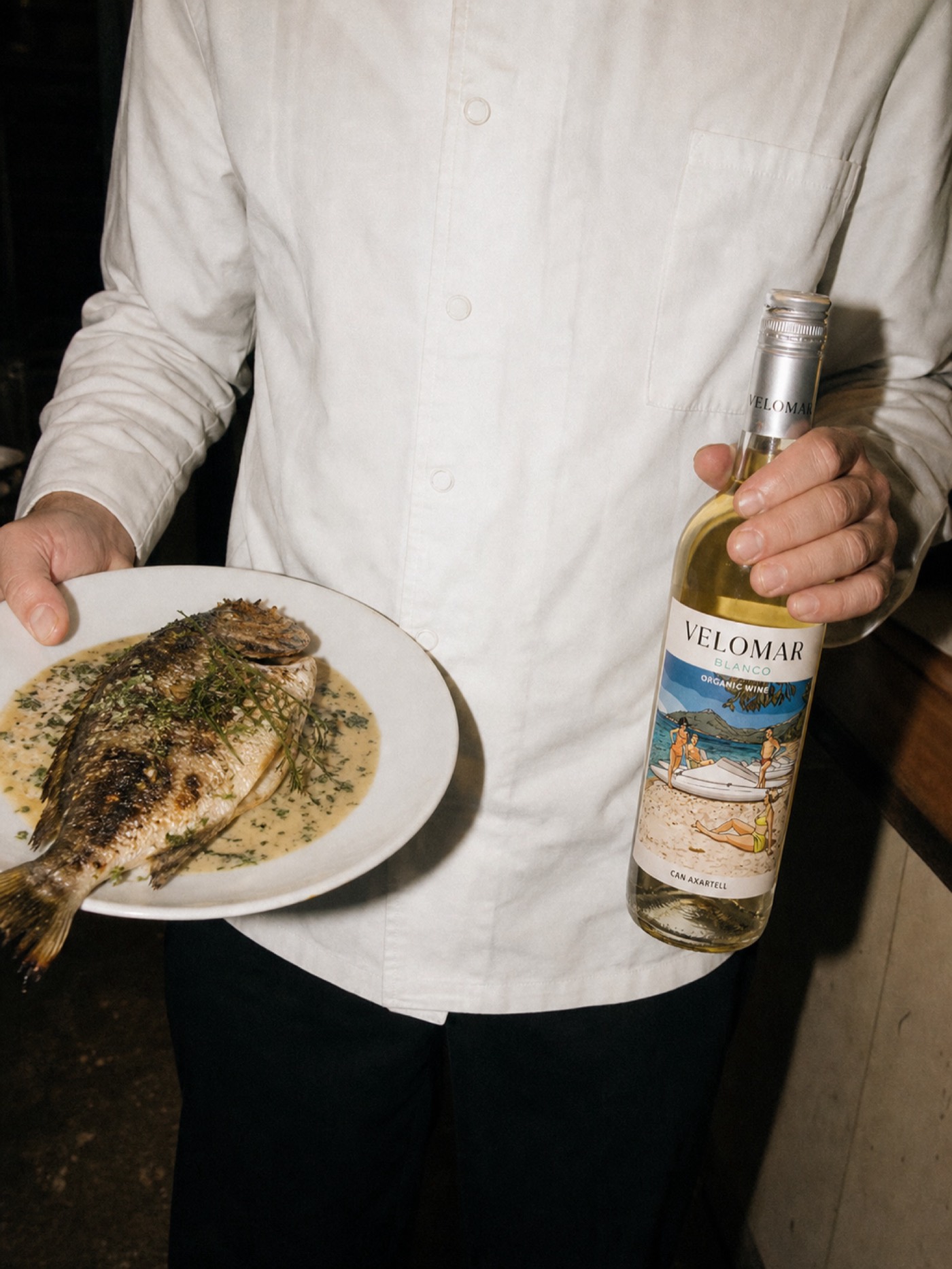 A chef holding a plate of grilled fish and a bottle of Velomar Blanco